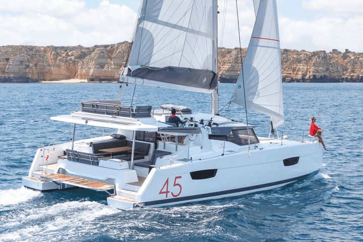 White sailing catamaran with sails open, number 45, sailing near rocky coast.