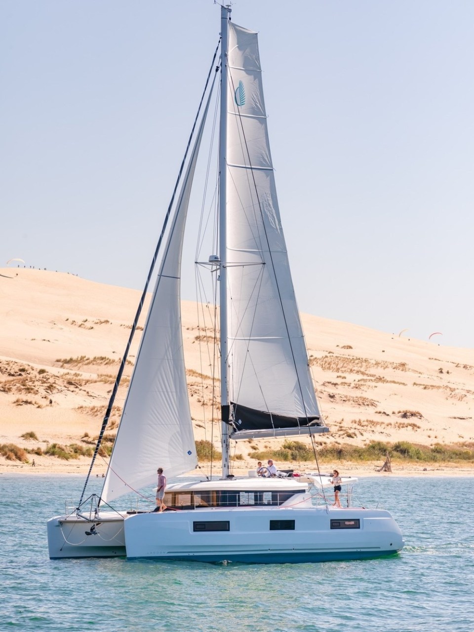 Catamaran sailing on calm water with sandy dunes in the background.