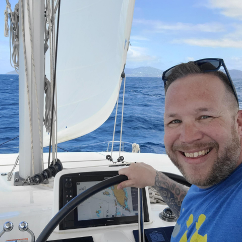 Man smiling while steering a sailboat in open ocean with a view of islands and blue skies.