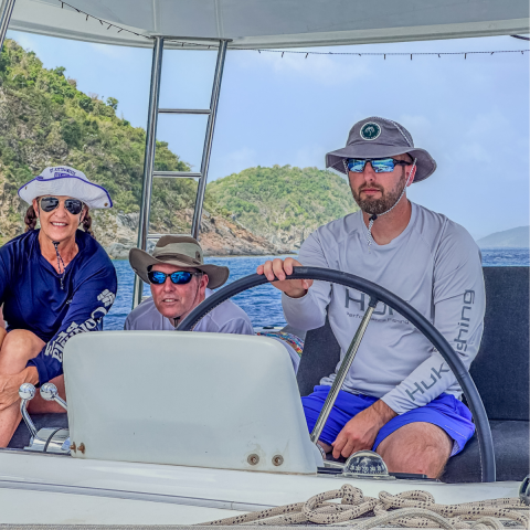 Three people on a boat, wearing hats and sunglasses, with a scenic island backdrop.